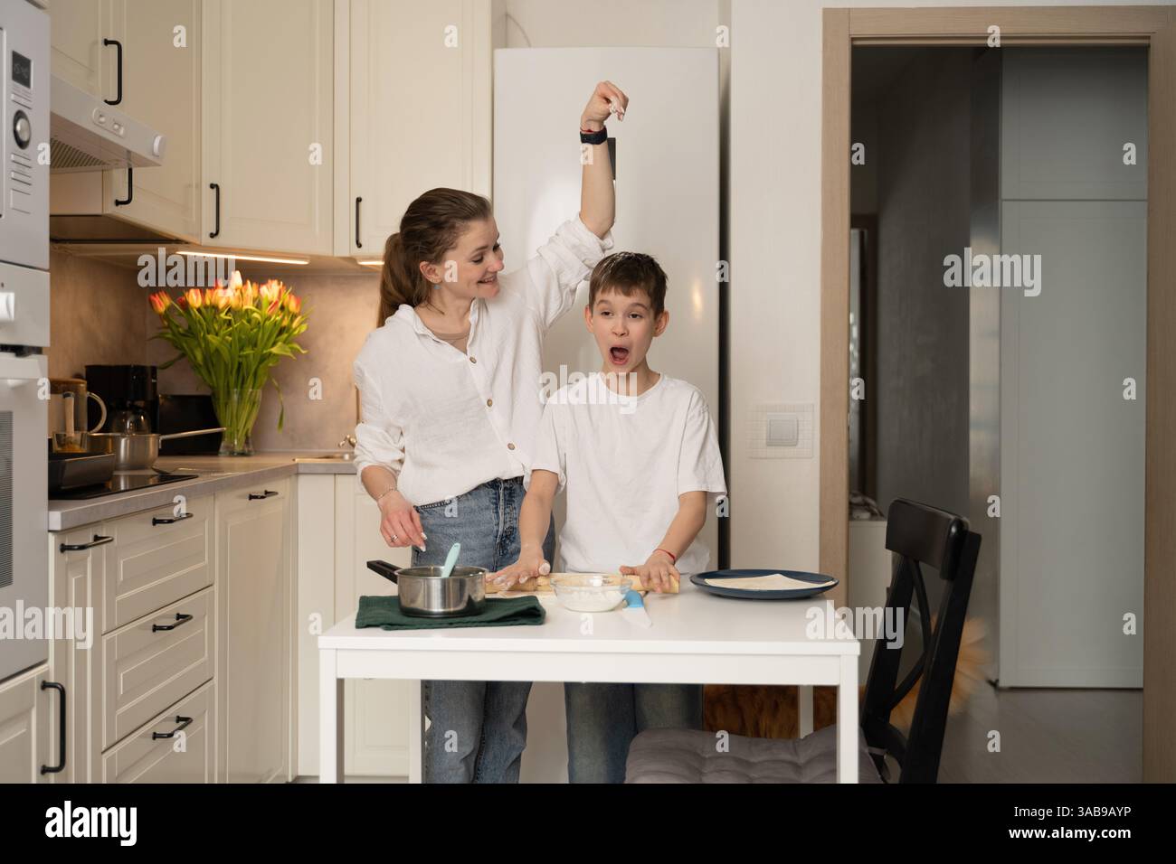 Una madre e un figlio gioiosi cuocono in una cucina moderna, condividendo un momento di divertimento. Il bambino sembra sorpreso mentre prepara gli ingredienti insieme, circondato da Foto Stock