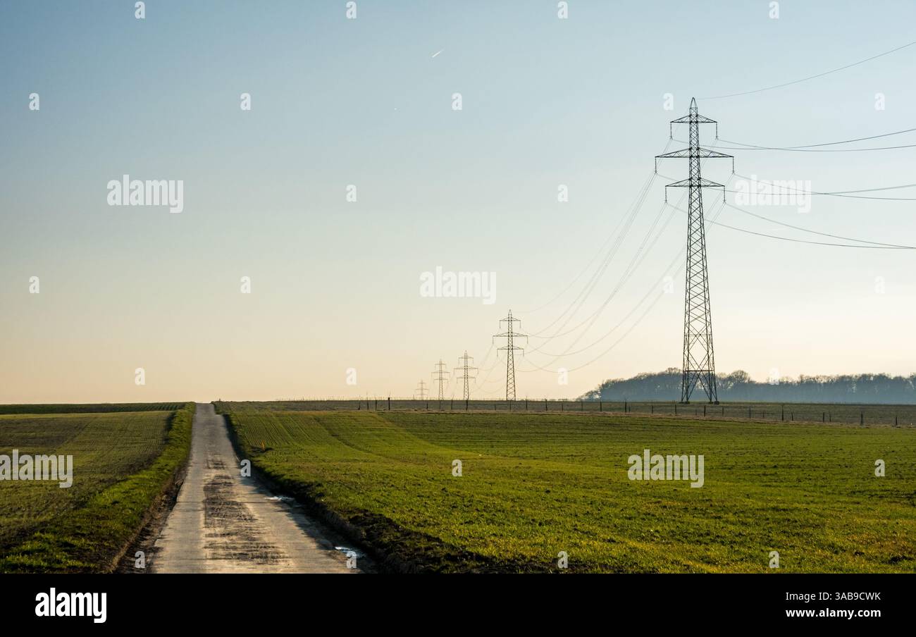 Un paesaggio rurale caratterizzato da una stretta strada che conduce attraverso campi verdi aperti, fiancheggiati da alte torri di trasmissione, adagiati su un cielo azzurro limpido con di Foto Stock