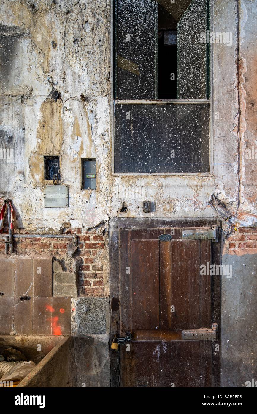 Un primo piano di una vecchia porta in legno posta all'interno di una parete in deterioramento con intonaco sbucciato, mattoni a vista e finestre invecchiate. Le trame evidenziano i pas Foto Stock