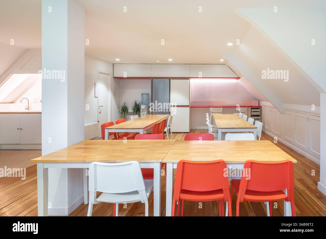 Luminosa cucina di coworking con sedie rosse e bianche, tavoli in legno e luce naturale. Design contemporaneo, ideale per riunioni e spazi collaborativi Foto Stock