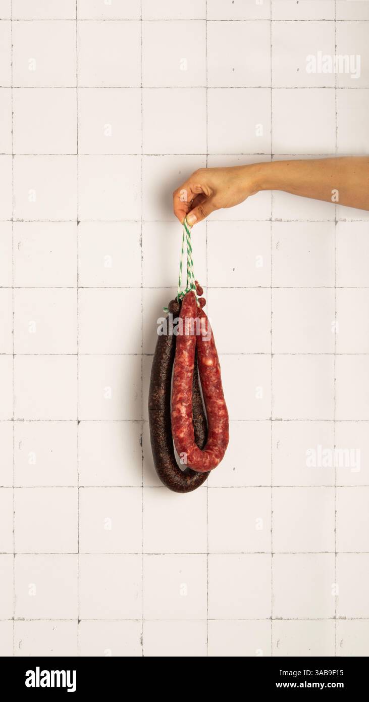 Una mano regge il budino nero e la salsiccia di maiale su una parete piastrellata bianca, mostrando texture e colori diversi. La composizione mette in risalto la cucina Foto Stock