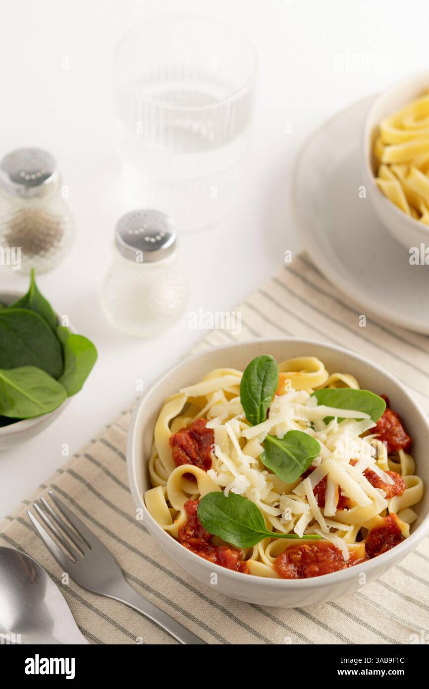Una ciotola di spaghetti condita con salsa di pomodoro, formaggio grattugiato e foglie di spinaci freschi. Accanto ad esso, un bicchiere d'acqua, sale e pepe shaker su uno stri Foto Stock