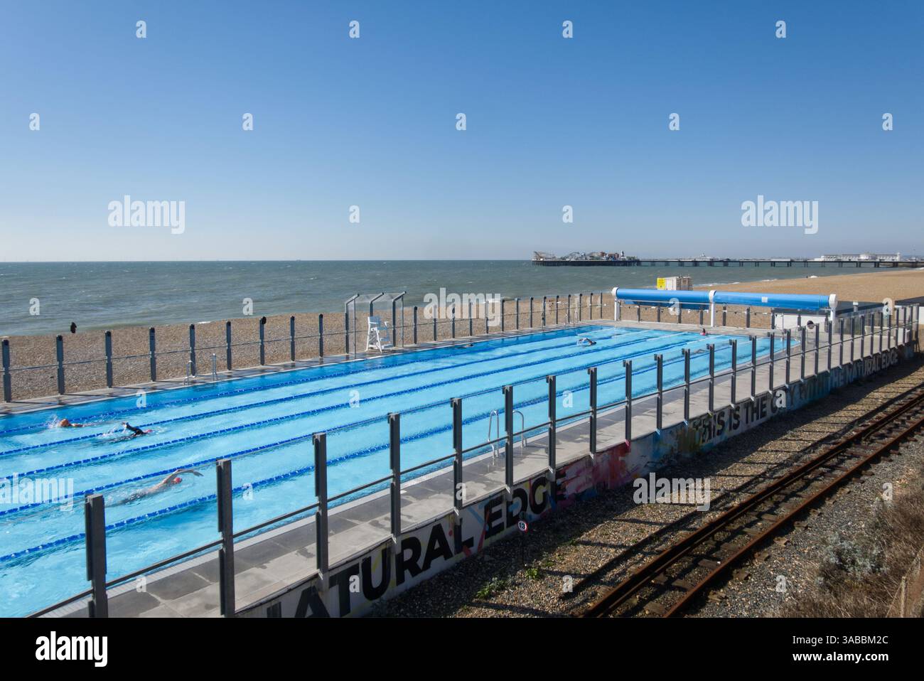 Piscina all'aperto sulla spiaggia di Brighton, East Sussex, Inghilterra. Con persone irriconoscibili che nuotano. Molo sullo sfondo. Linea ferroviaria in primo piano. Foto Stock