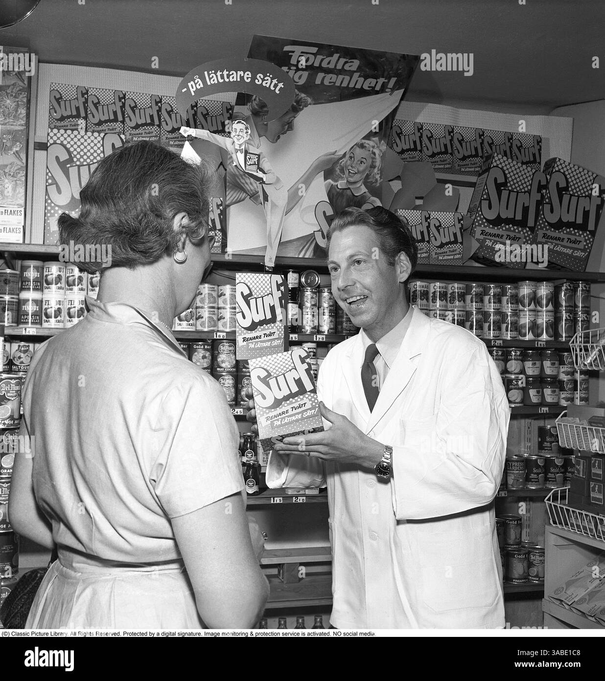 Un classico negozio degli anni '1950, in cui un commesso, vestito con un cappotto bianco, presenta con entusiasmo il nuovo detergente Surf al cliente. Sorreggendo la confezione, sembra che ne stia spiegando i vantaggi, evidenziando la sua eccellente capacità di pulizia e la sua praticità. La cliente donna ascolta attentamente mentre l'impiegato mette in mostra il prodotto. Gli scaffali dei negozi sullo sfondo sono riforniti di vari prodotti in scatola e di articoli confezionati, riflettendo lo stile ben organizzato dei negozi di alimentari della metà del secolo. Le pubblicità per il detergente Surf sono esposte in modo prominente sulle pareti, con slogan Foto Stock
