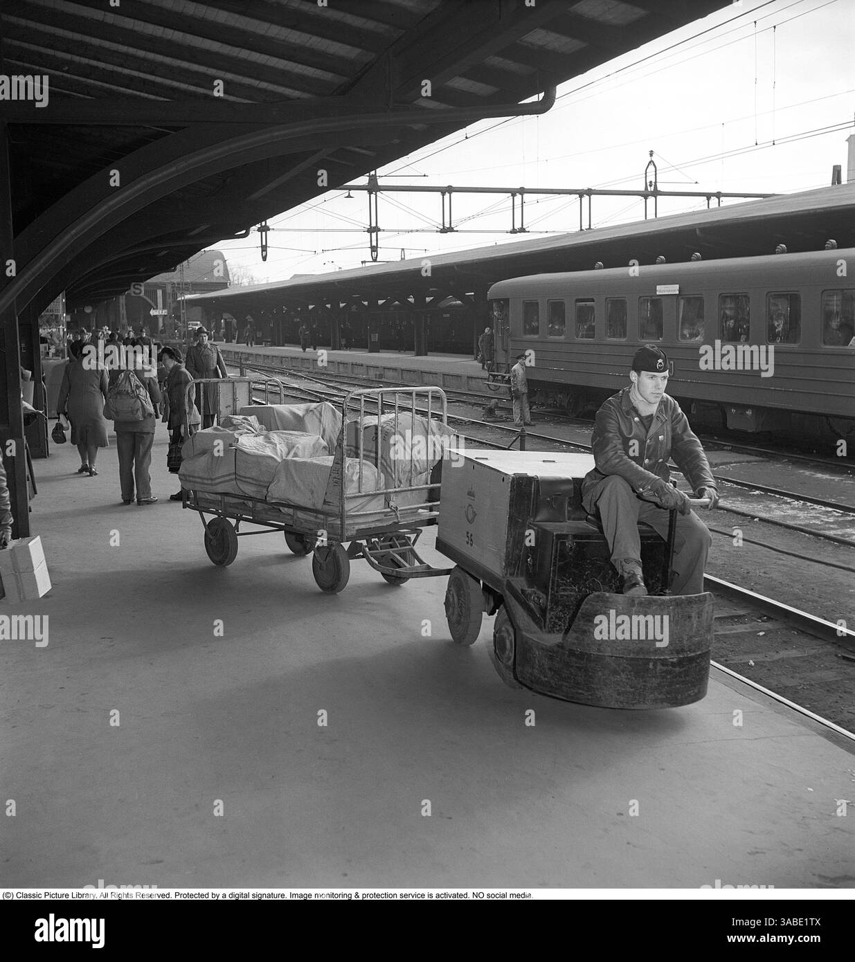 Una stazione ferroviaria degli anni '1950 Sulla piattaforma, un uomo sta guidando un piccolo veicolo elettrico utilizzato per il trasporto di lettere e pacchi per posta. Al veicolo è collegato un carrello. La piattaforma è protetta da un'ampia struttura del tetto, che fornisce copertura ai passeggeri e ai lavoratori. Una panoramica di come la logistica ferroviaria e le operazioni quotidiane delle stazioni ferroviarie apparivano negli anni '1950, mostrando l'uso di veicoli elettrici e il vivace movimento di persone e merci tipico dell'epoca. 1955 Foto Stock