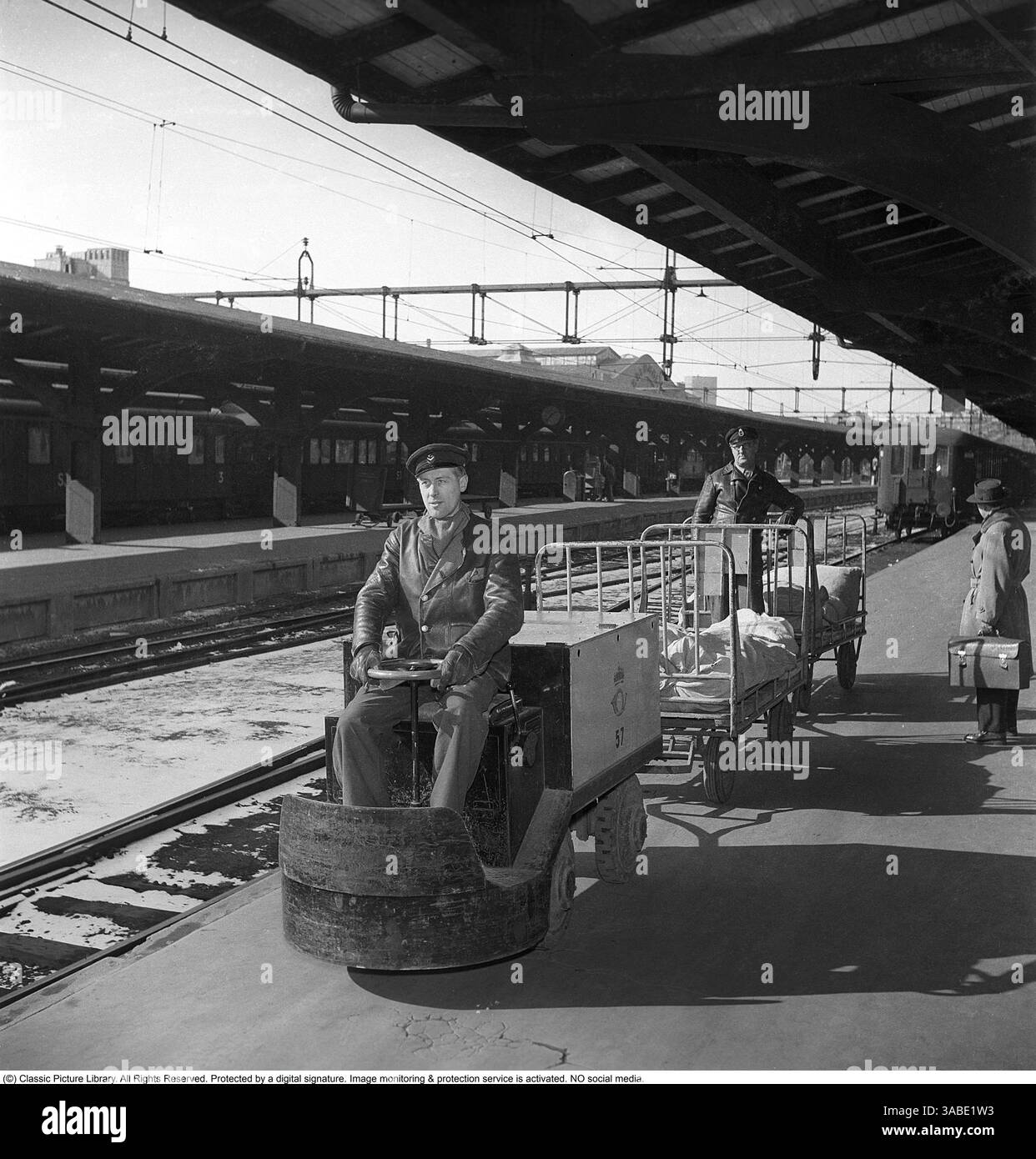 Una stazione ferroviaria degli anni '1950 Sulla piattaforma, un uomo sta guidando un piccolo veicolo elettrico utilizzato per il trasporto di lettere e pacchi per posta. Attaccato al veicolo c'è un carrello con un altro uomo in piedi in cima al carrello, apparentemente supervisionando il trasporto. A destra, una persona cammina lungo la piattaforma, portando con sé una valigetta, forse sulla strada per prendere un treno. Sullo sfondo, i vagoni ferroviari sono visibili lungo i cavi elettrici sospesi, a indicare che la stazione ospita treni elettrici. La piattaforma è protetta da un'ampia struttura del tetto, che fornisce copertura ai passeggeri e. Foto Stock