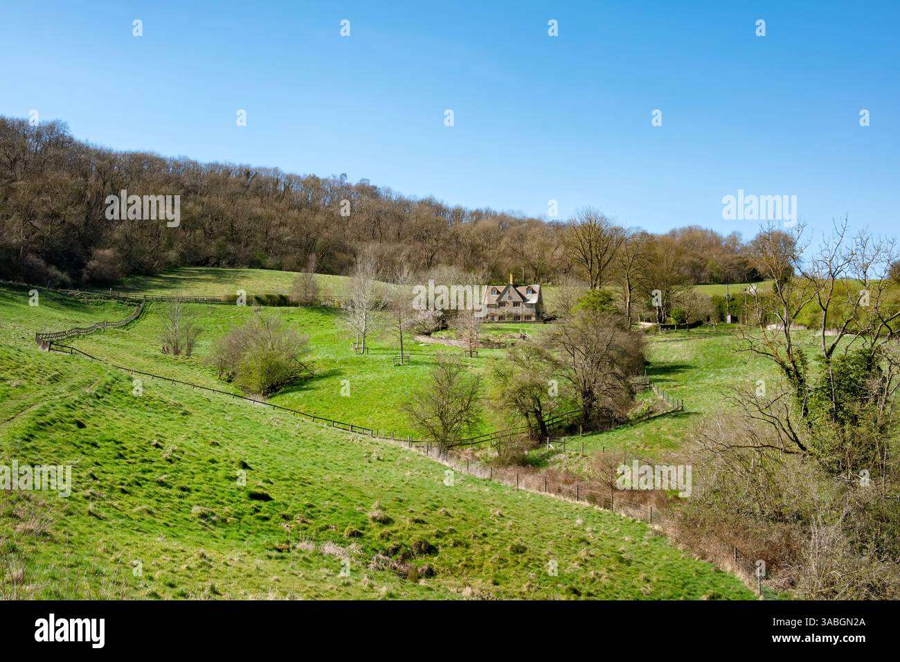 Cotswolds, Gloucestershire. Una vista sulla campagna verde aperta verso una tradizionale fattoria in pietra annidata sotto un orizzonte alberato Foto Stock