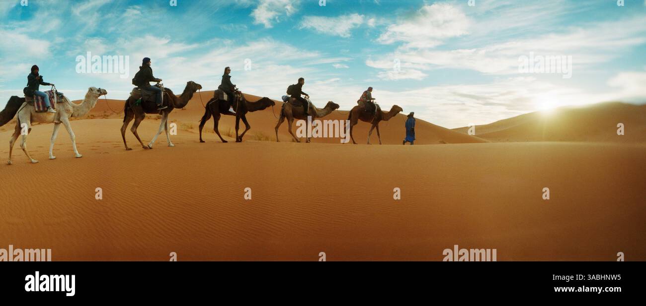 Vista panoramica dei turisti che cavalcano cammelli attraverso il paesaggio del deserto del Sahara condotto da un uomo berbero, il Marocco Foto Stock