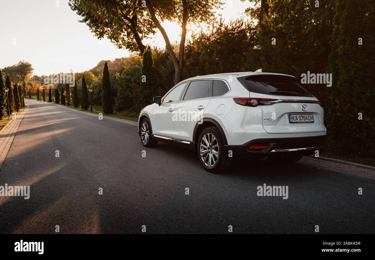 Mazda CX-9 bianca su una strada pavimentata di campagna costeggiata da cipressi. Vista posteriore grandangolare di tre quarti del SUV di famiglia al tramonto. Foto Stock