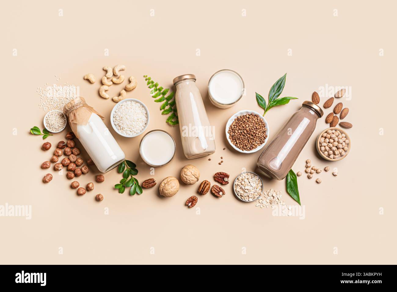 Vari flaconi di latte a base di piante vegane, ingredienti e foglie verdi. Latte senza lattosio, senza lattosio con noci e grani su sfondo beige, creativo Foto Stock