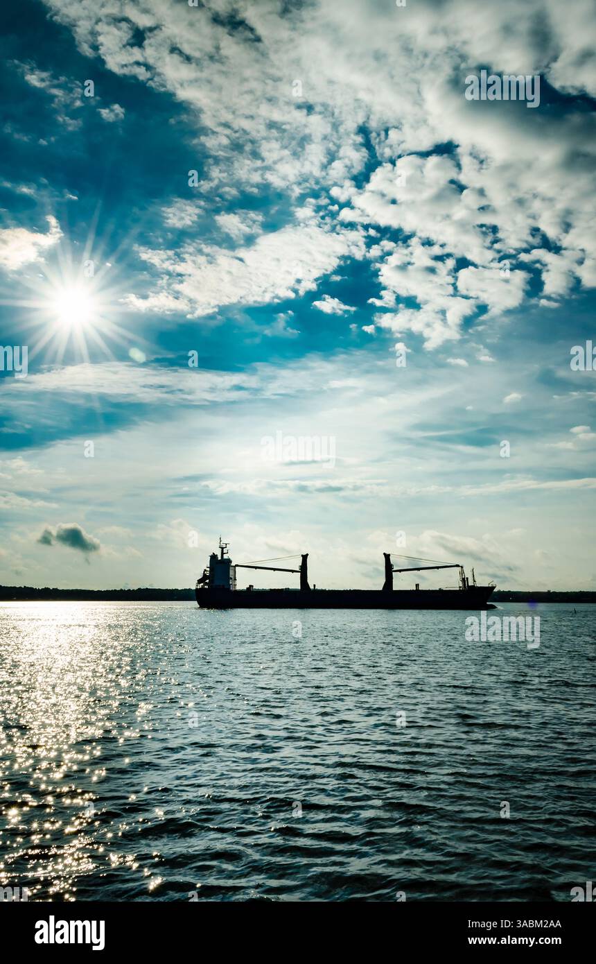 La nave da carico si staglia con un sole stellato dietro di essa contro un cielo blu nuvoloso e un'acqua blu scuro Foto Stock
