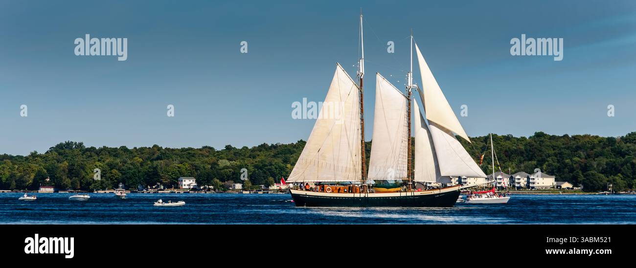 Panorama di una nave alta in navigazione completa lungo il fiume St. Lawrence accanto a imbarcazioni più piccole con la costa dello stato di New York e cottage in distanc Foto Stock