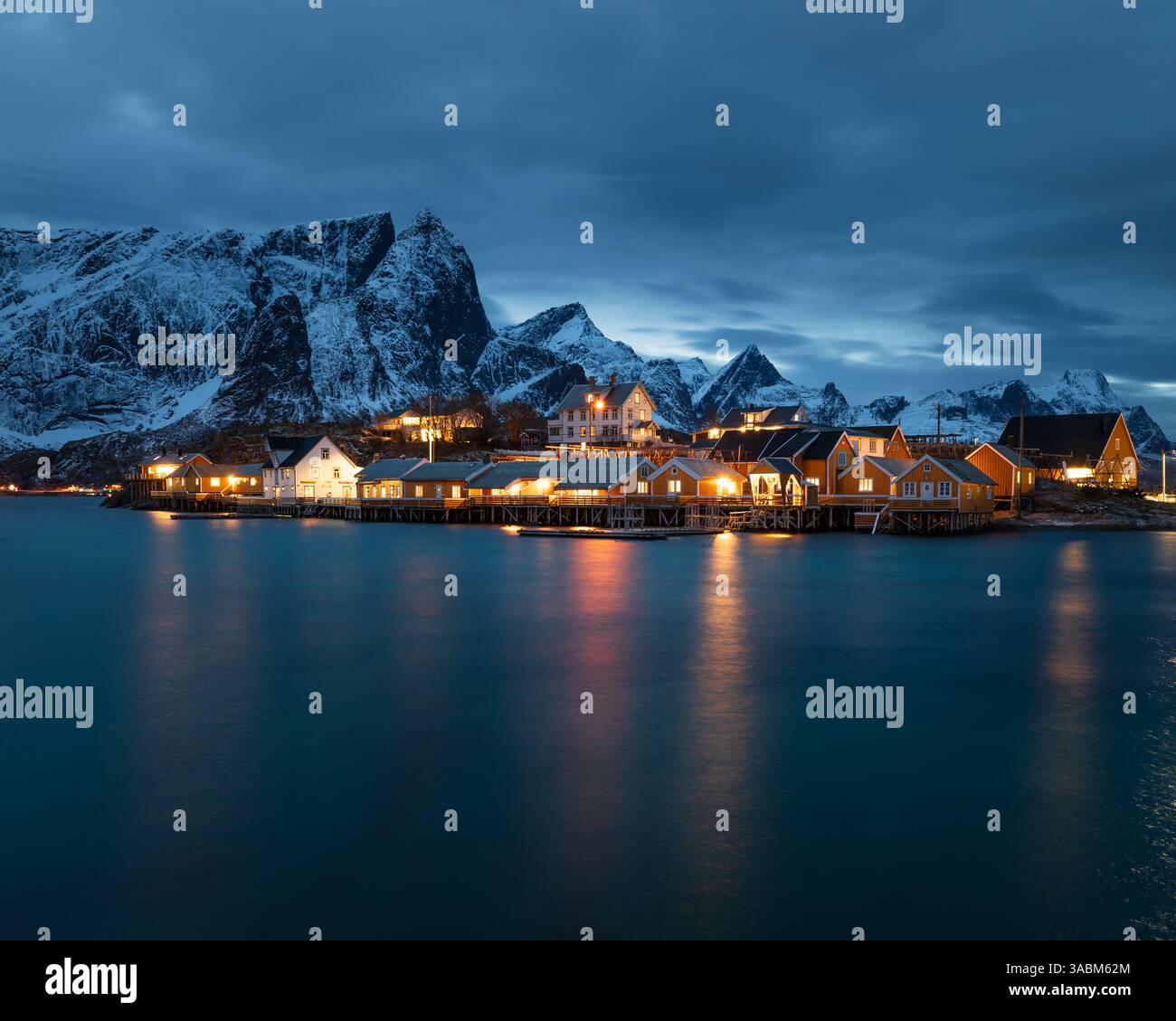 Sakrisoy auf den Lofoten in Norwegen zur blauen Stunde mit beleuchteten gelben Rorbuer am Fjord und Spiegelung im stillen Wasser unter dunklem Himmel. Foto Stock