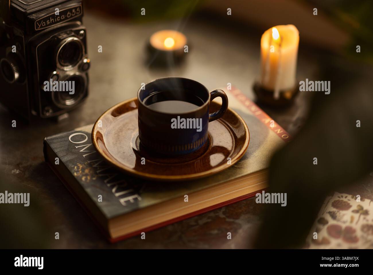 Primo piano di una tazza di caffè caldo blu vintage in piedi su un vecchio libro in un davanzale in pietra circondato da candele. Foto Stock