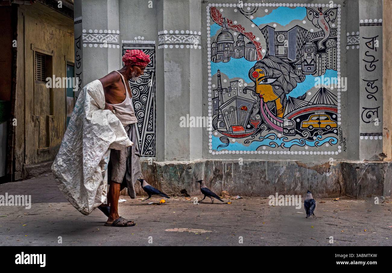 Un anziano rapinatore cammina davanti a un murale colorato su una parete di strada di Kolkata, riflettendo i contrasti di vita, arte e India urbana. Foto Stock