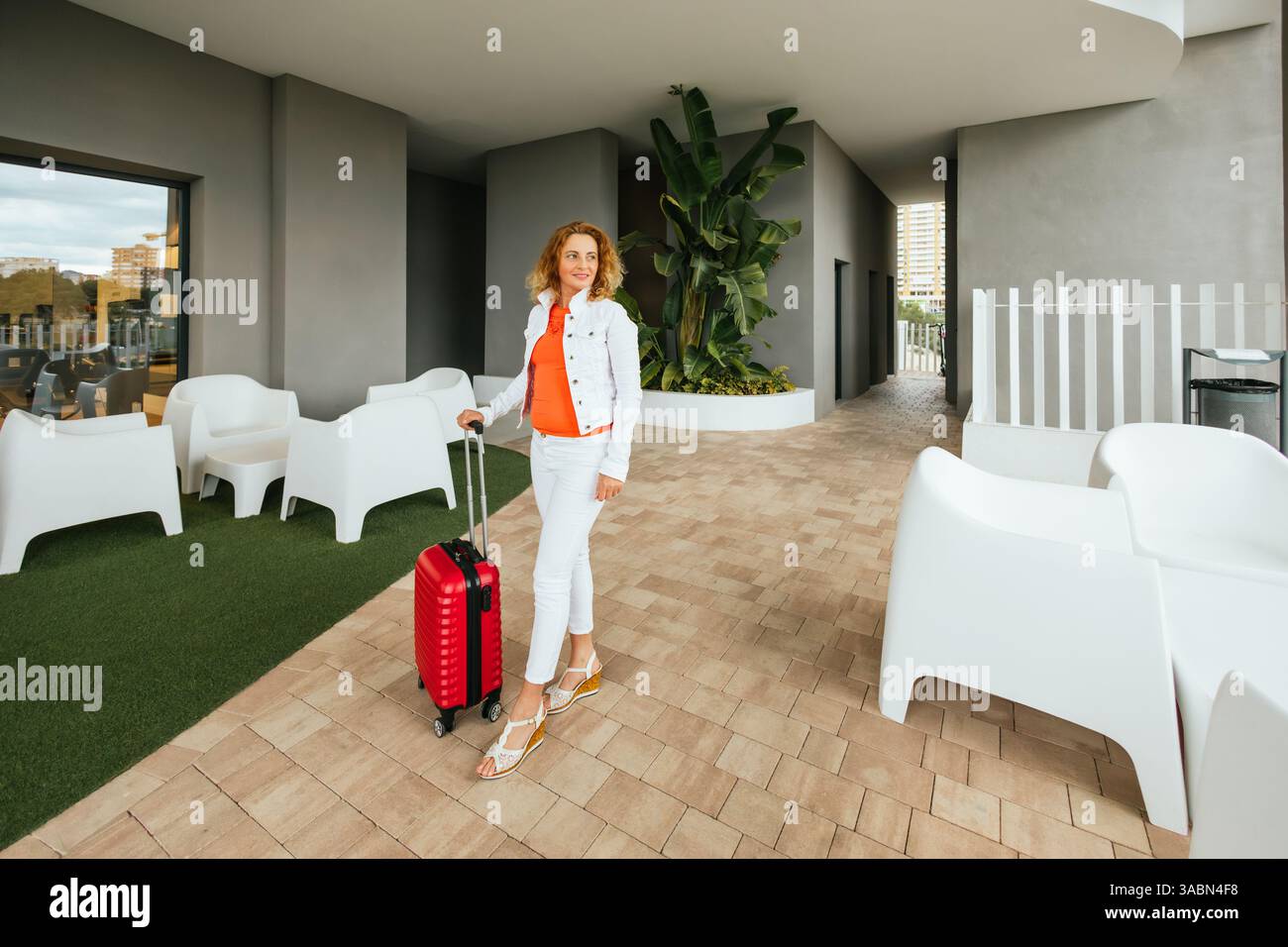 Donna di mezza età in abiti bianchi e arancioni che arriva in un hotel contemporaneo con una valigia rossa, in piedi vicino a mobili moderni e piante tropicali Foto Stock
