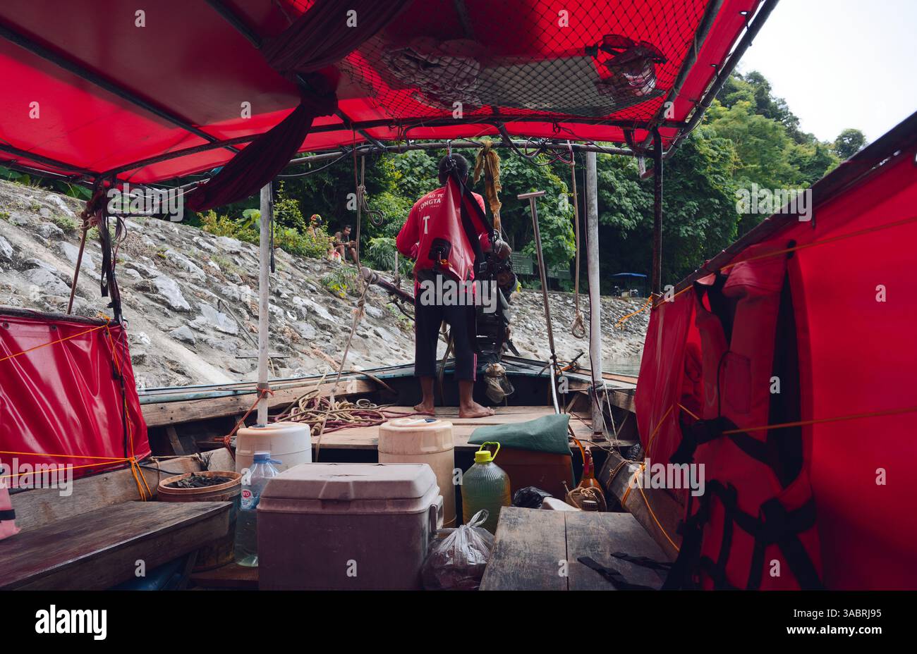 Il capitano della barca a coda lunga si prepara ad attraccare sotto i vivaci teloni rossi vicino a una costa rocciosa artificiale Foto Stock