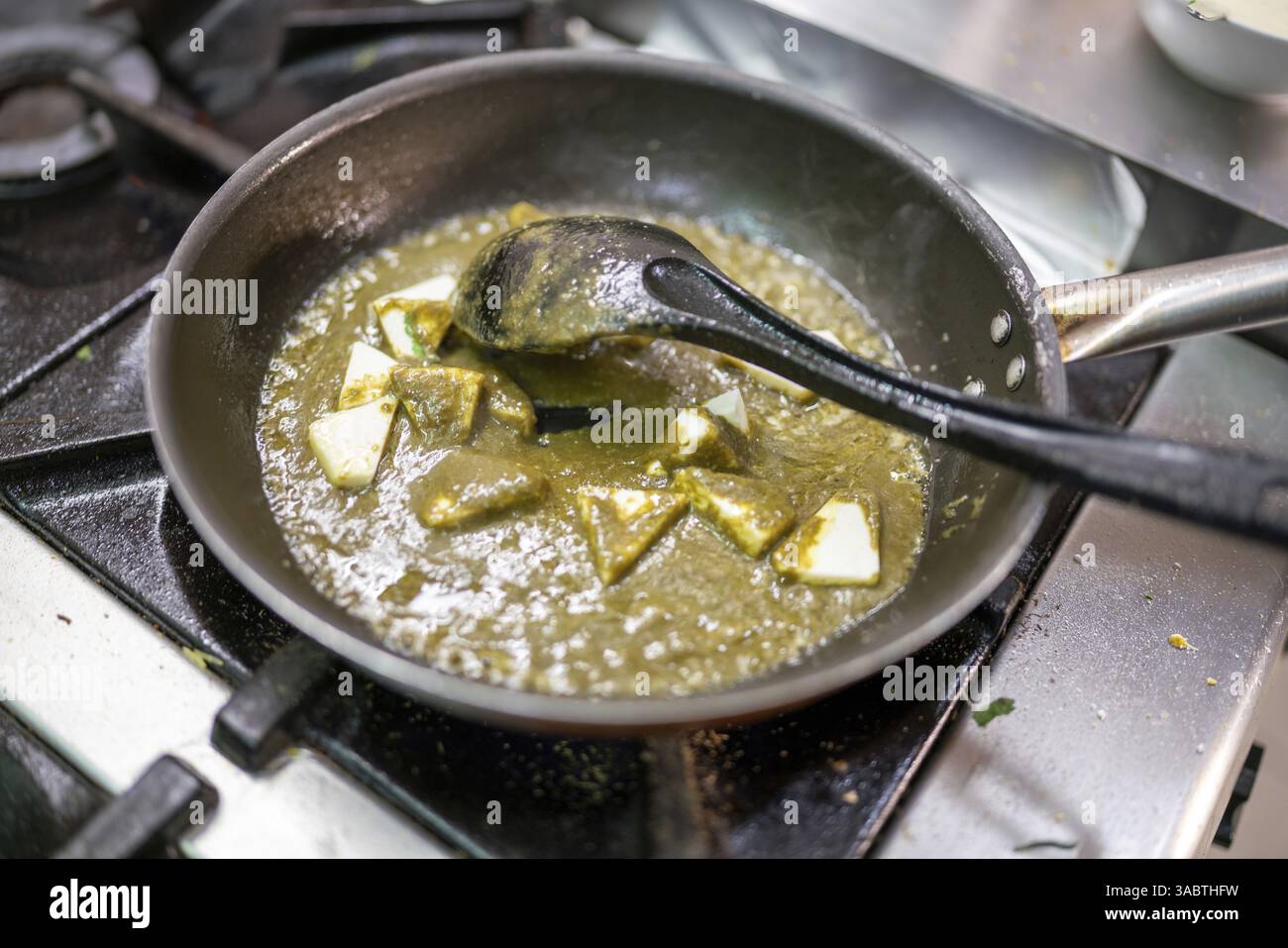 Lo chef professionista mescola il paneer di palak, un popolare piatto indiano a base di spinaci e formaggio, utilizzando un cucchiaio nero in una cucina professionale Foto Stock