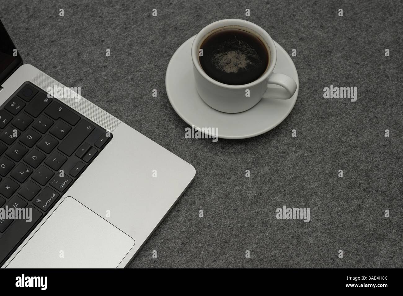 Deutschland, 03.04.2025, primo piano di un computer portatile moderno accanto a una tazza bianca di caffè nero su una superficie in feltro grigio. L'ambiente suggerisce un bancomat di lavoro tranquillo Foto Stock