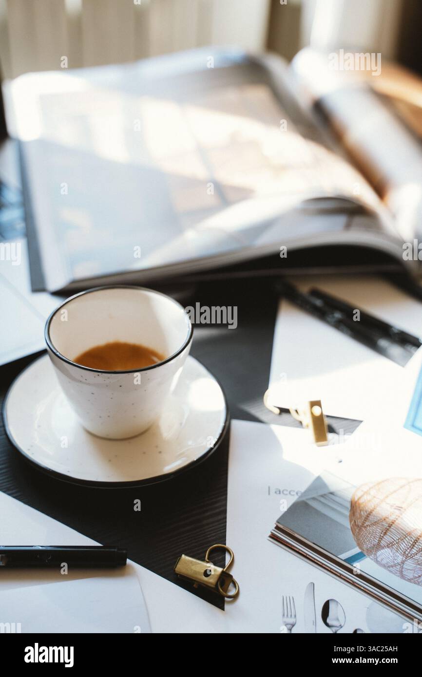 Una tazza di caffè siede su una scrivania circondata da riviste, giornali e cancelleria, creando un'atmosfera accogliente e produttiva in un ambiente di lavoro al sole naturale Foto Stock