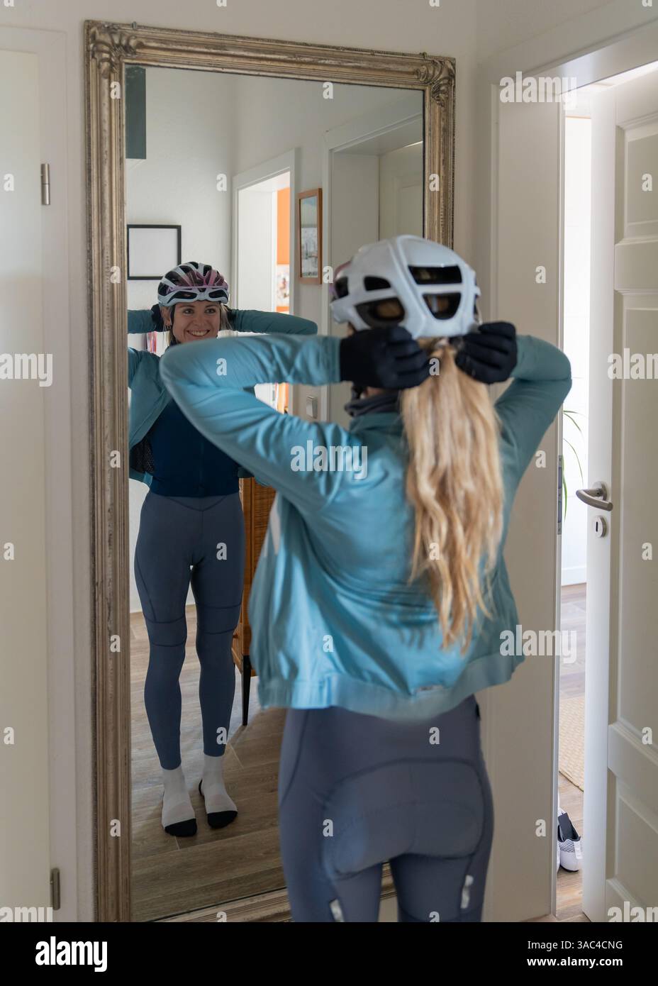 Una giovane ciclista atletica in uniforme da ciclismo e casco si trova davanti allo specchio, fissando i capelli prima di un allenamento. L'immagine atmosferica trasmette il momento o. Foto Stock