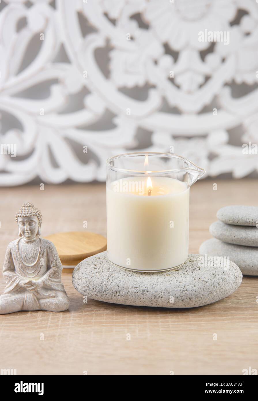Candela profumata fatta in casa con olio per massaggi in tazza di vetro con beccuccio. Sfondo in stile boemo, statuetta di Buddha seduta, pietre zen impilate per rilassarsi. Foto Stock