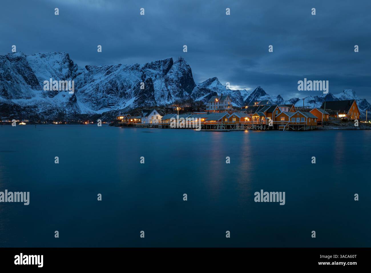Sakrisoy auf den Lofoten in Norwegen zur blauen Stunde mit beleuchteten gelben Rorbuer am Fjord und Spiegelung im stillen Wasser unter dunklem Himmel. Foto Stock