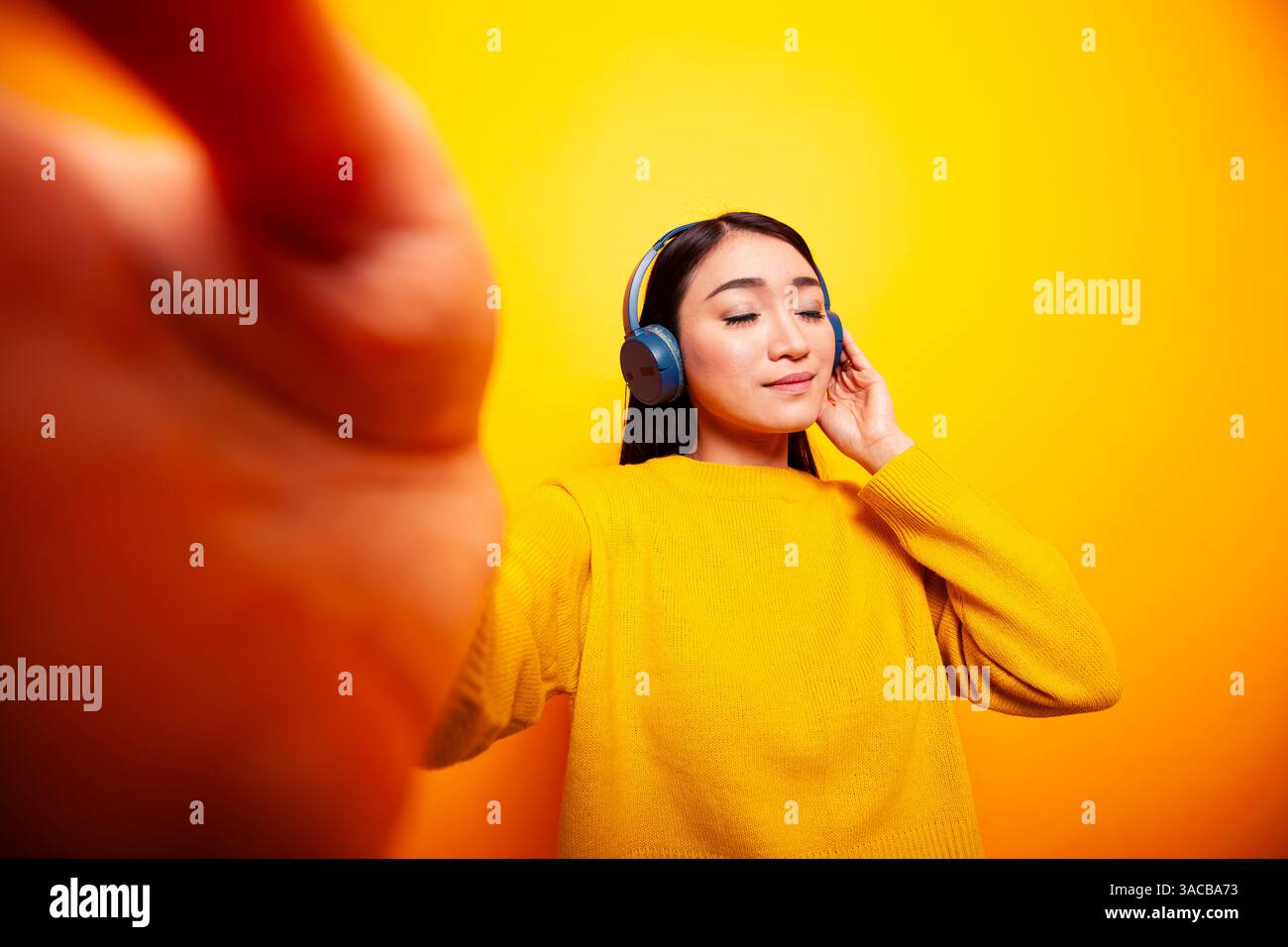 Vlogger asiatico in piedi su sfondo isolato e ascoltare musica. Giovane donna con le cuffie, che registra il vlog quotidiano e tiene in mano una fotocamera, con un'espressione rilassata davanti allo sfondo giallo. Foto Stock