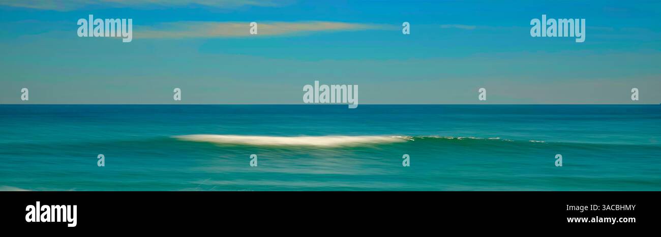 Una tranquilla scena di un'onda dolce che si infrangono su una spiaggia sabbiosa mentre il sole tramonta, che proietta un caldo bagliore sull'acqua e sulla sabbia. L'acqua è un turchese vibrante Foto Stock
