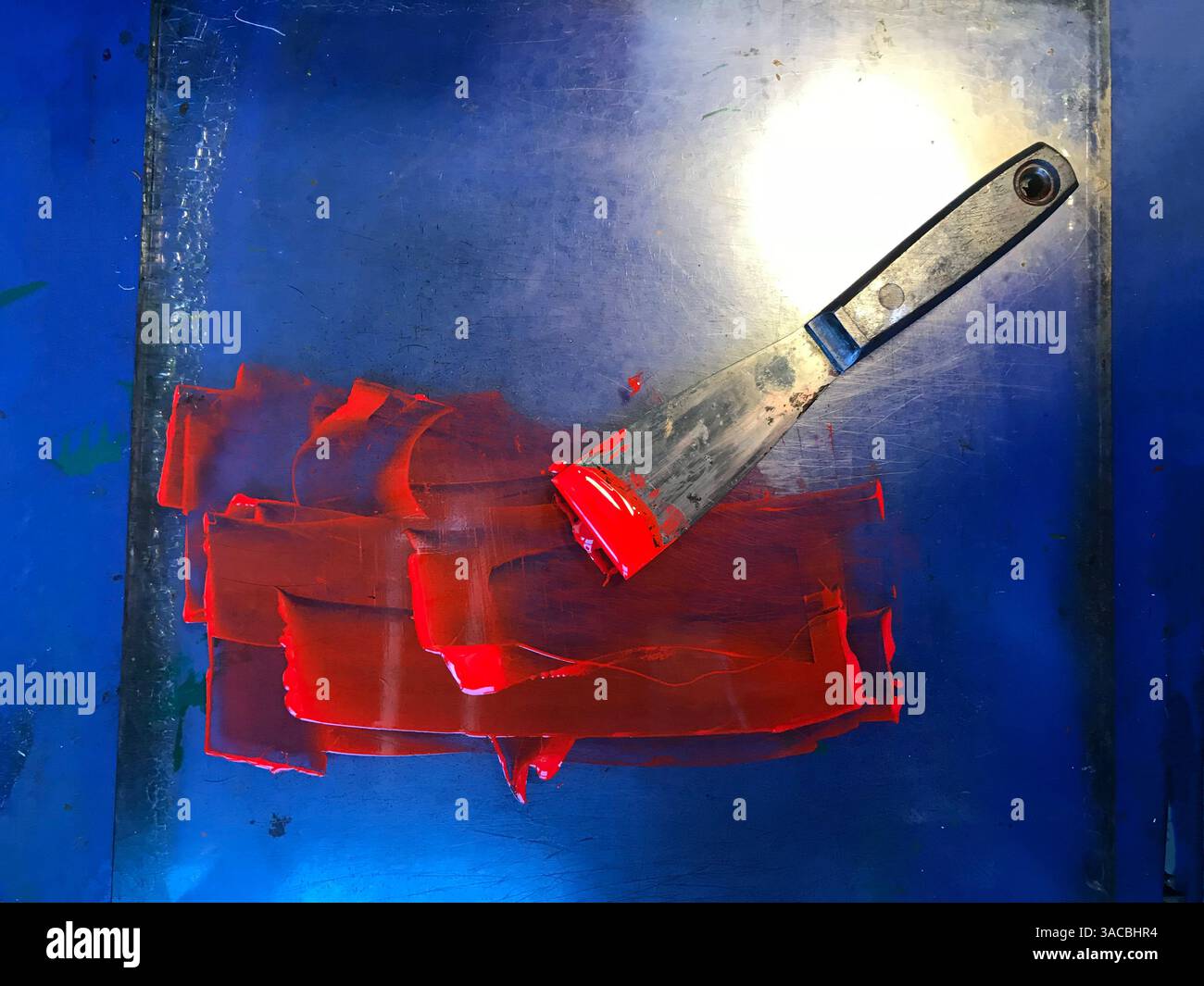 L'inchiostro rosso brillante viene distribuito su una superficie blu con una spatola metallica che poggia accanto, per mostrare un momento creativo nello studio di un artista, Rev Foto Stock