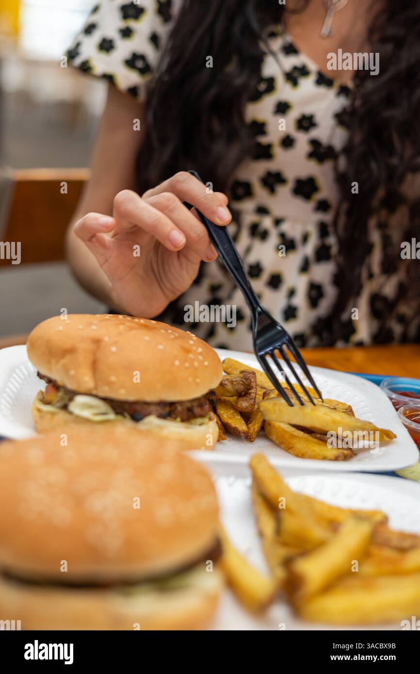 Una donna mangia patatine fritte usando una forchetta al tavolo di un ristorante, con il suo hamburger nelle vicinanze, mescolando la cena informale con una presentazione ordinata. Foto Stock