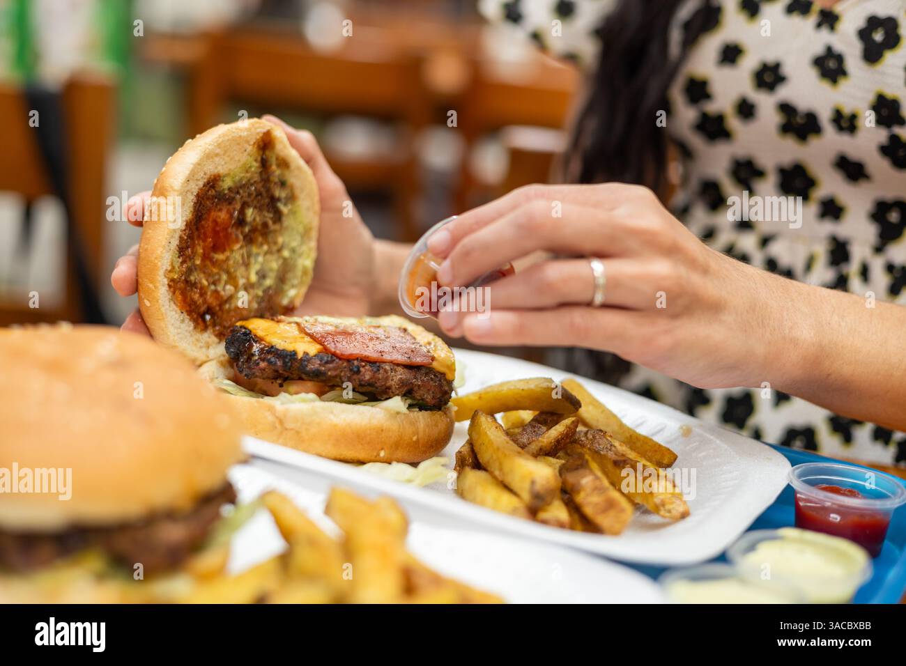 Una donna prepara il suo hamburger con salsa, circondato da patatine fritte in un ambiente informale pieno di sapore e anticipazione. Foto Stock