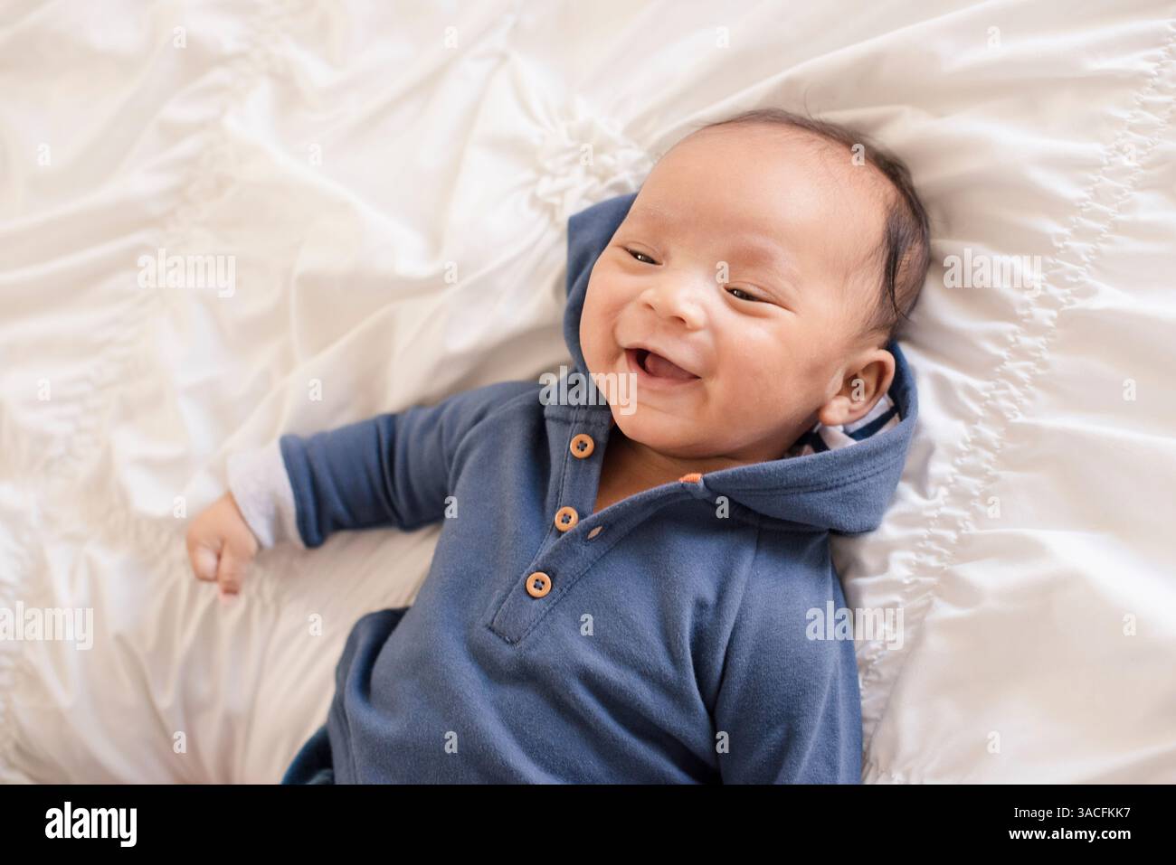 Bambino sorridente disteso su soffici letti bianchi con cappuccio blu Foto Stock