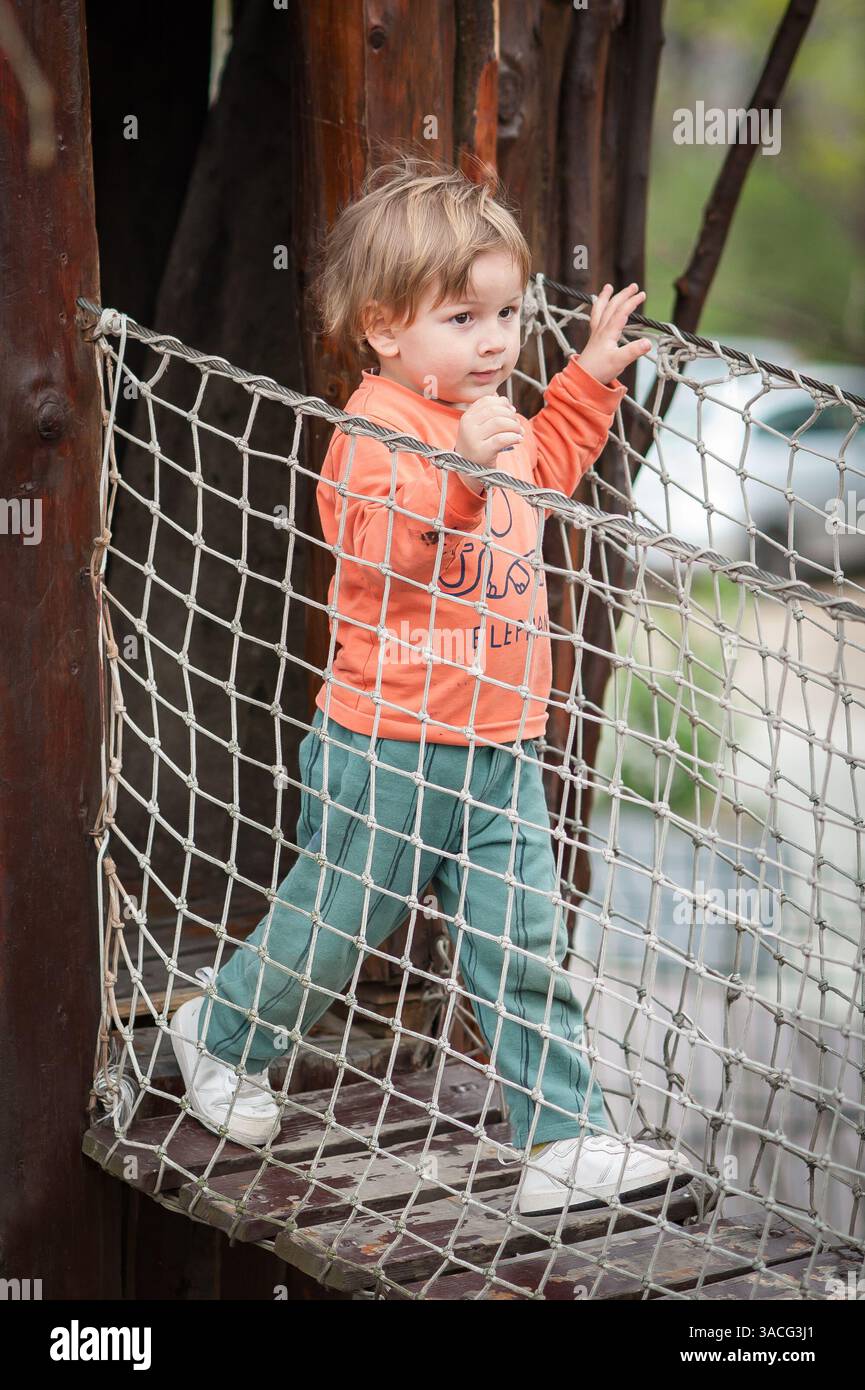 Un bambino con una camicia arancione e pantaloni verdi naviga con attenzione su una rete di corda in un'area di gioco naturale, divertendosi all'aperto in un parco Foto Stock