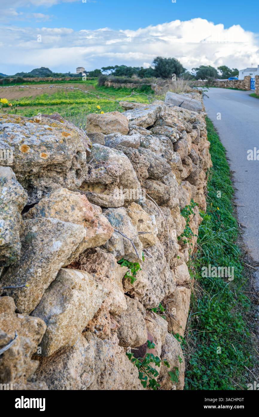 Un rustico muro di pietra fiancheggia una tranquilla strada rurale a Minorca, catturando l'essenza del tranquillo fascino della campagna dell'isola. Foto Stock