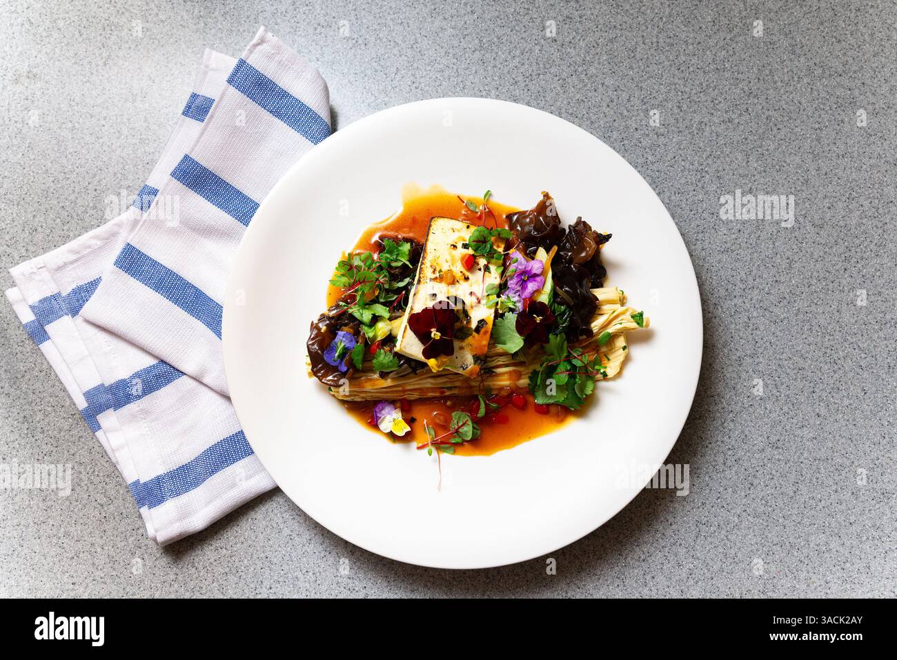 Pasto gourmet tofu placcato con funghi auricolari in legno, microgreen, fiori commestibili e salsa vivace servita su un piatto bianco. Foto Stock