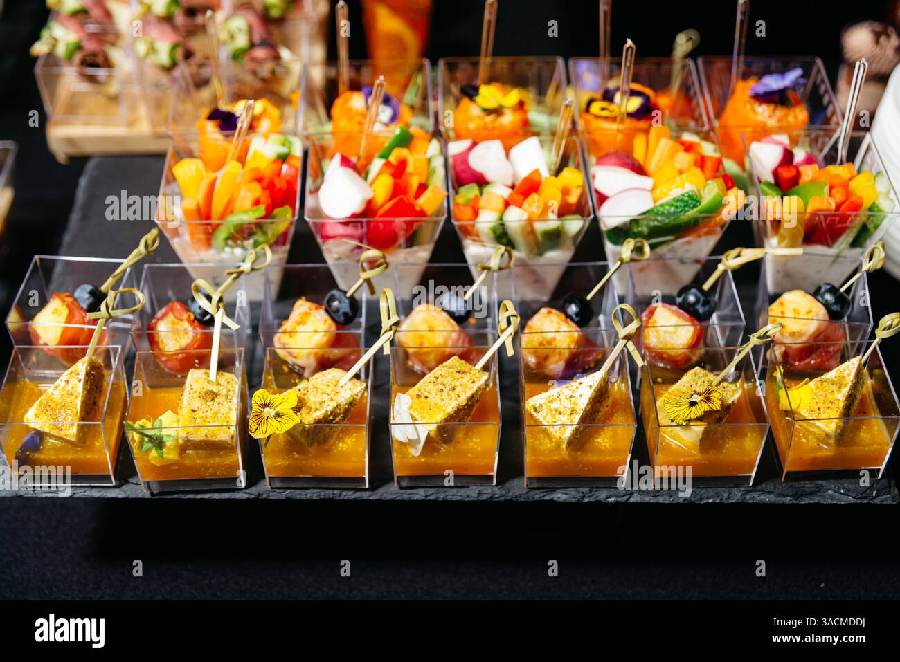 Una varietà di eleganti antipasti serviti in bicchieri di plastica trasparente, con verdure, formaggi, frutta e fiori commestibili per eventi. Foto Stock