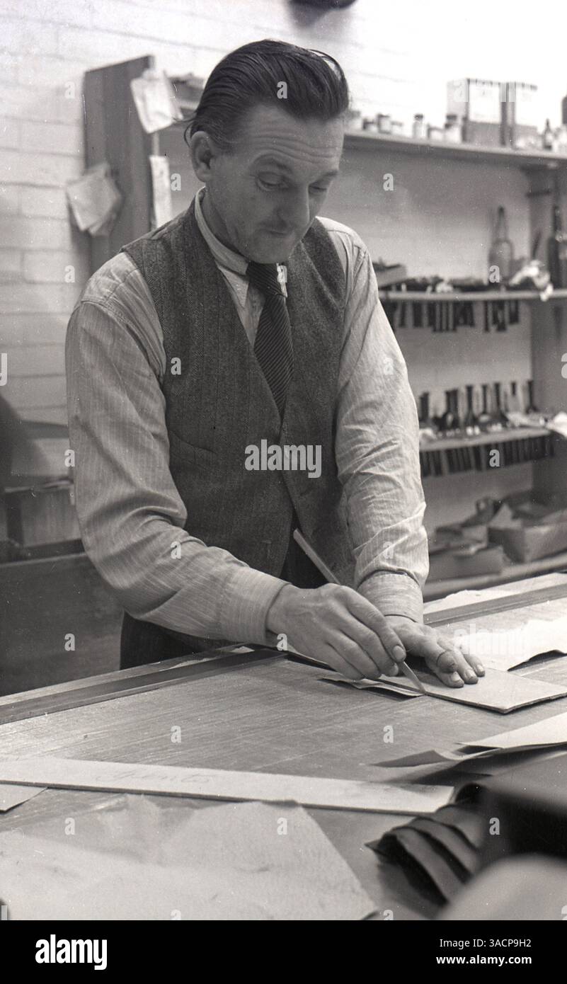 1930, storico, un uomo adulto con maglietta e cravatta e gilet che lavora all'interno di un'officina in piedi su una panchina usando un bisturi per tagliare il cartoncino. Foto Stock