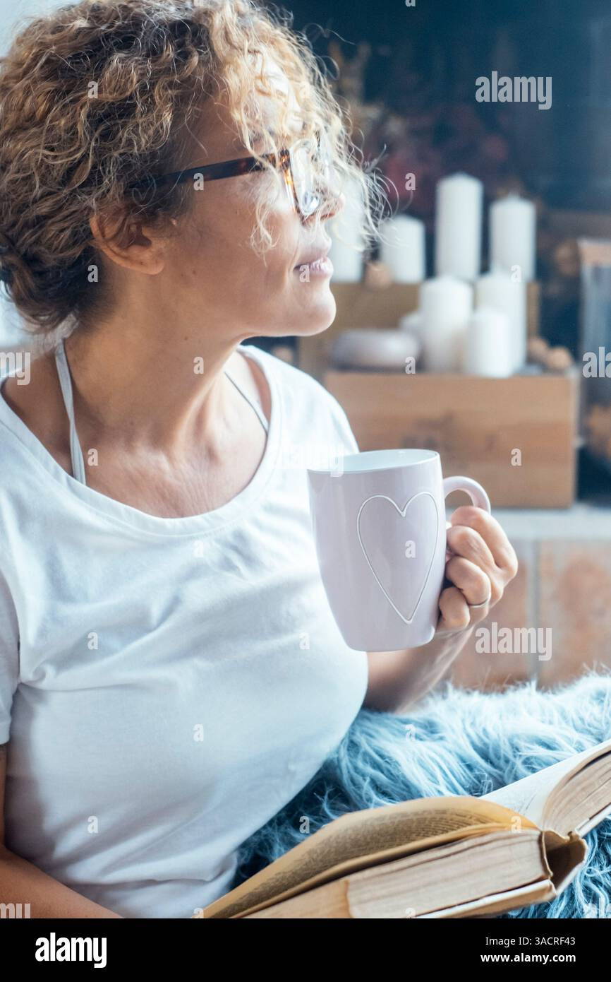 Una donna adulta che si rilassa a casa con una tazza calda di tè o caffè in mano, sembra serena e romantica, con un'espressione pacifica, godendosi il suo tempo personale in un ambiente accogliente e tranquillo Foto Stock