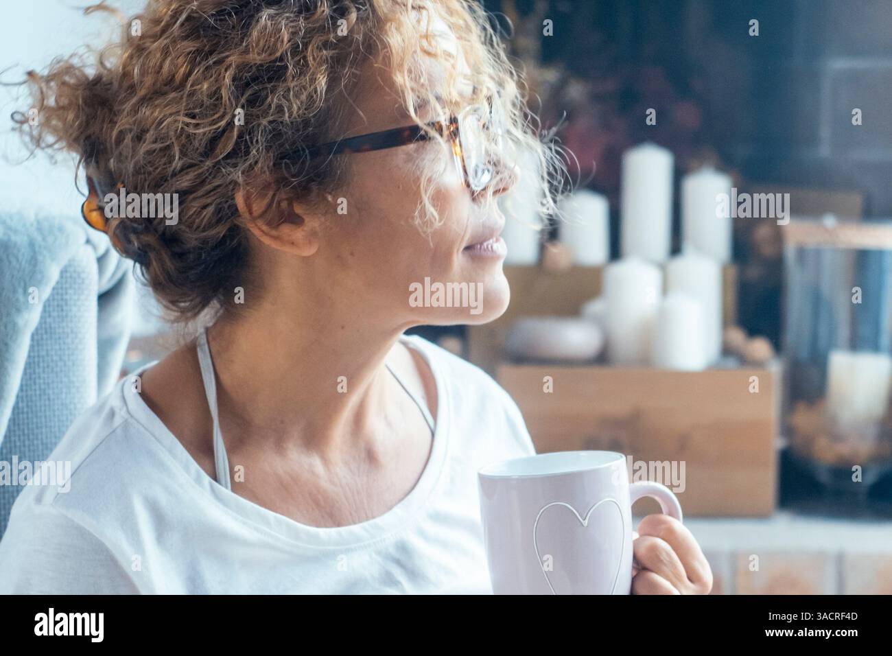 Una donna adulta che si rilassa a casa con una tazza calda di tè o caffè in mano, sembra serena e romantica, con un'espressione pacifica, godendosi il suo tempo personale in un ambiente accogliente e tranquillo Foto Stock