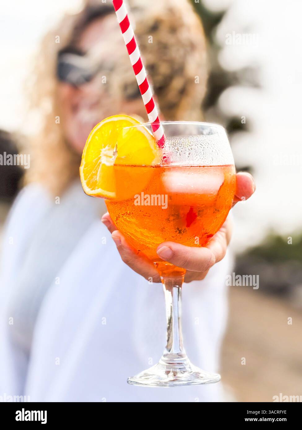Un'elegante scena estiva con una donna sorridente che offre un cocktail di spritz arancio con cubetti di ghiaccio e paglia, in un'atmosfera luminosa e vivace, perfetta per socializzare Foto Stock