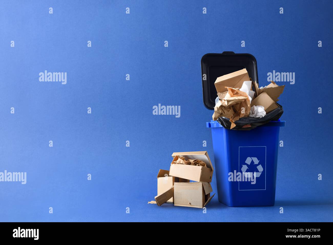 Contenitore blu dei rifiuti con simbolo di riciclaggio riempito con cartone e scarti di carta e altre scatole e carta accanto a esso, isolata con Foto Stock