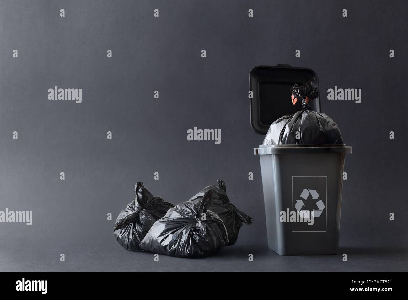 Contenitore per rifiuti grigio con simbolo di riciclaggio riempito con sacchetto per rifiuti e altri sacchetti accanto a esso sul terreno isolato con sfondo grigio a gradiente. Fo Foto Stock