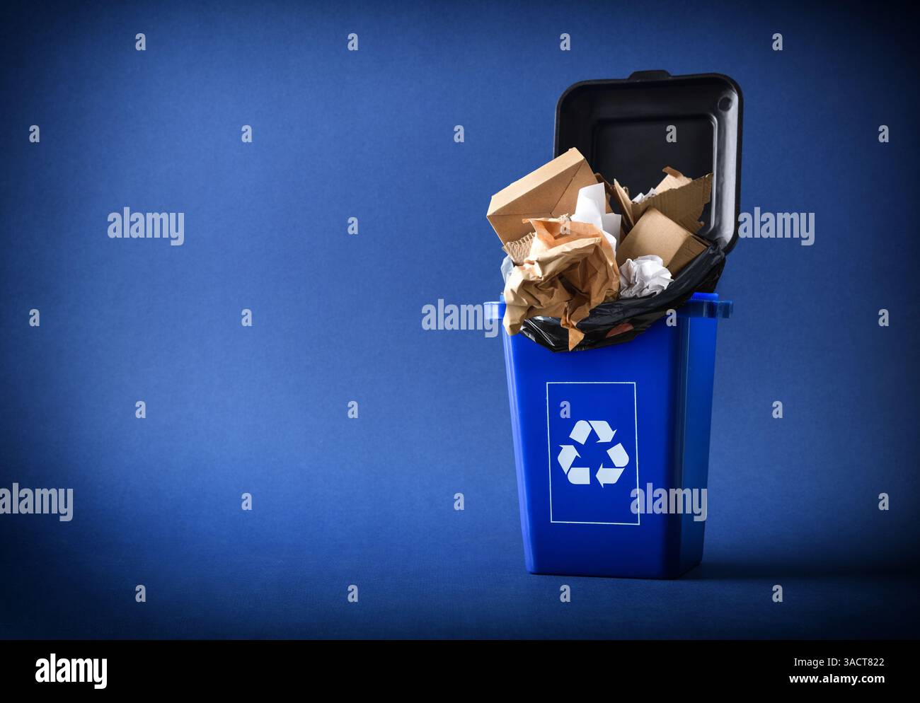 Concetto di riciclaggio di cartone e carta con un contenitore blu con il simbolo di riciclaggio e un sacchetto nero isolato su sfondo blu. Vista frontale. Foto Stock