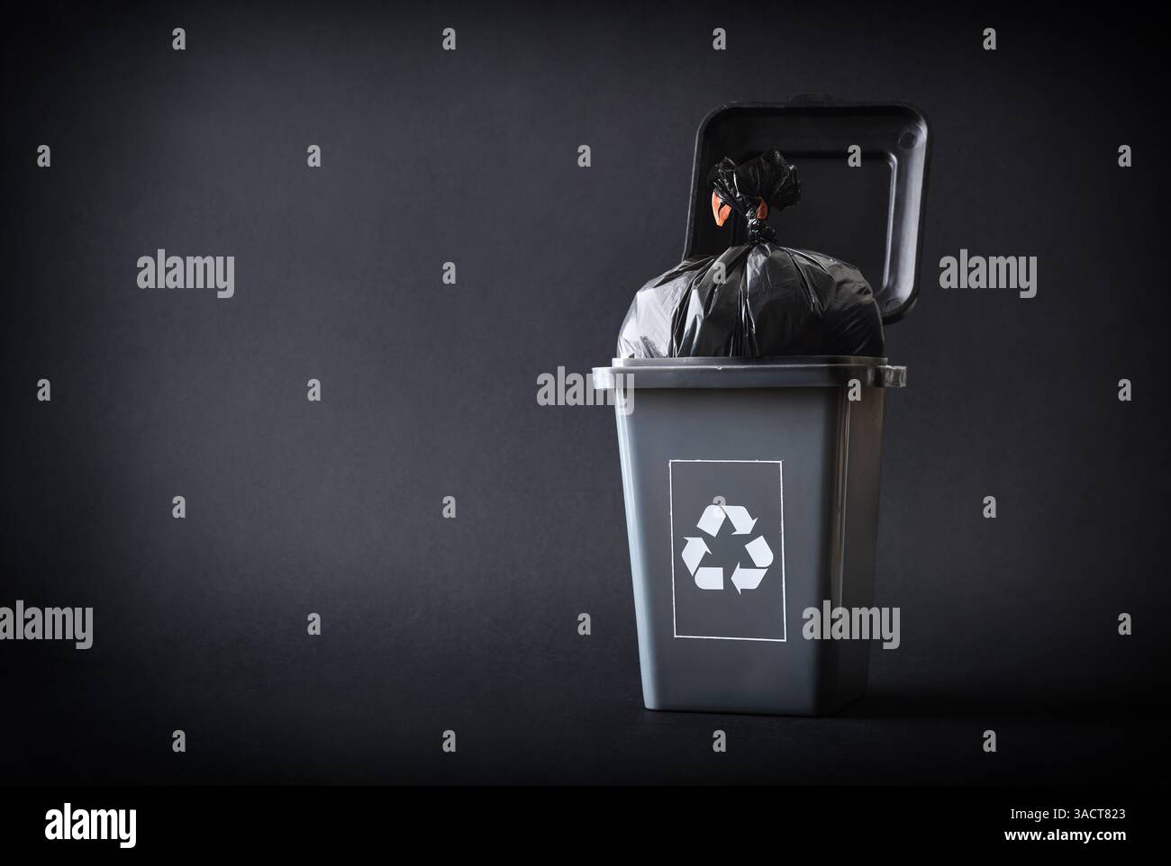 Concetto generico di riciclaggio dei rifiuti con un contenitore di riciclaggio grigio con simbolo di riciclaggio riempito con un sacchetto nero isolato su sfondo grigio. Vista frontale. Foto Stock
