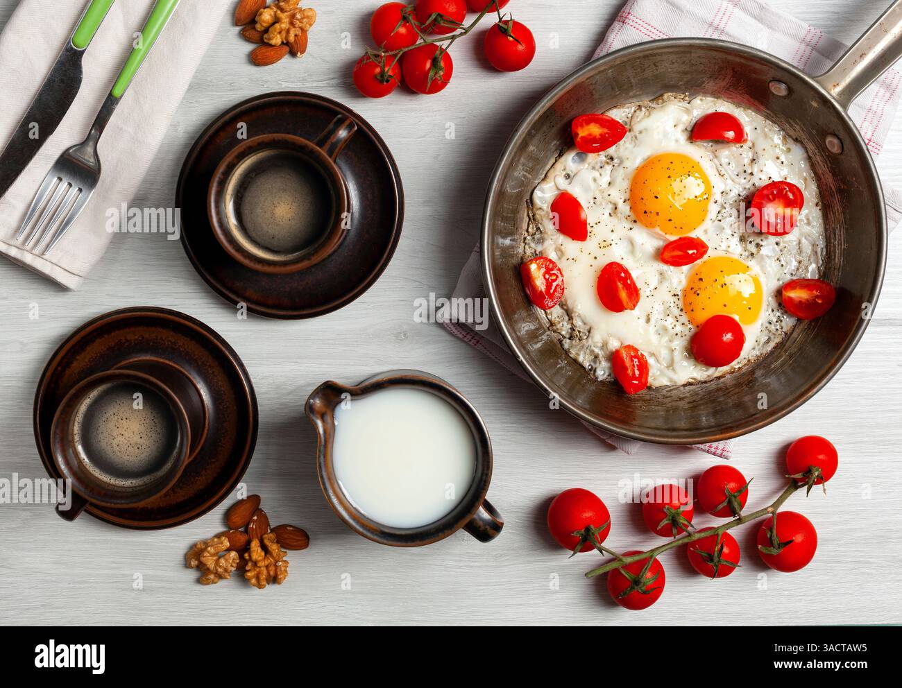 Due uova fritte con pomodori ciliegini in una padella, due tazze di caffè, latte, noci e rami di pomodori ciliegini stanno componendo una deliziosa colazione su un tavolo di legno bianco Foto Stock