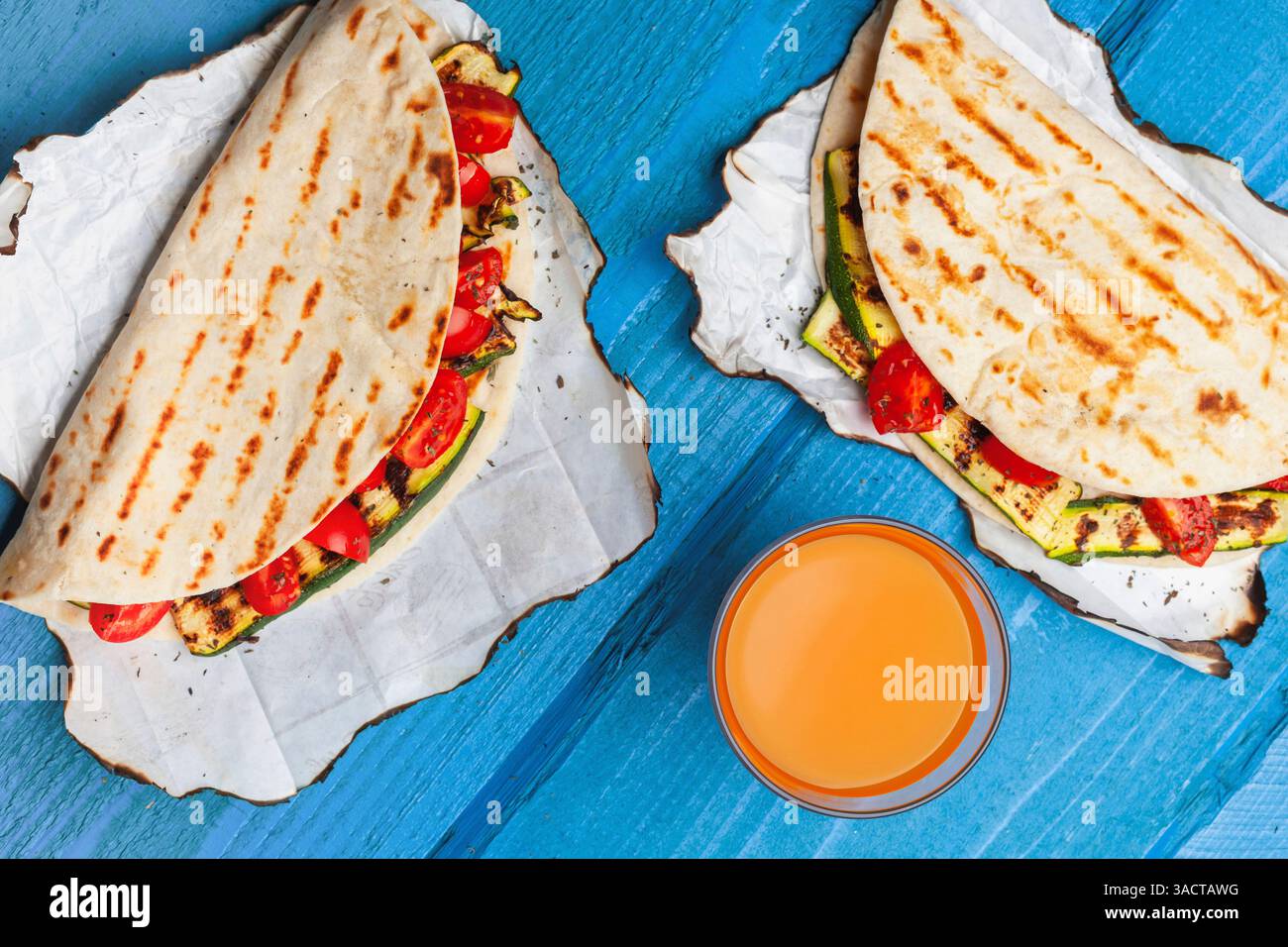 Due piadine vegane con zucchine grigliate e pomodori ciliegini serviti su carta da forno con un bicchiere di succo d'arancia su un tavolo di legno blu, per un pasto sano e rinfrescante Foto Stock
