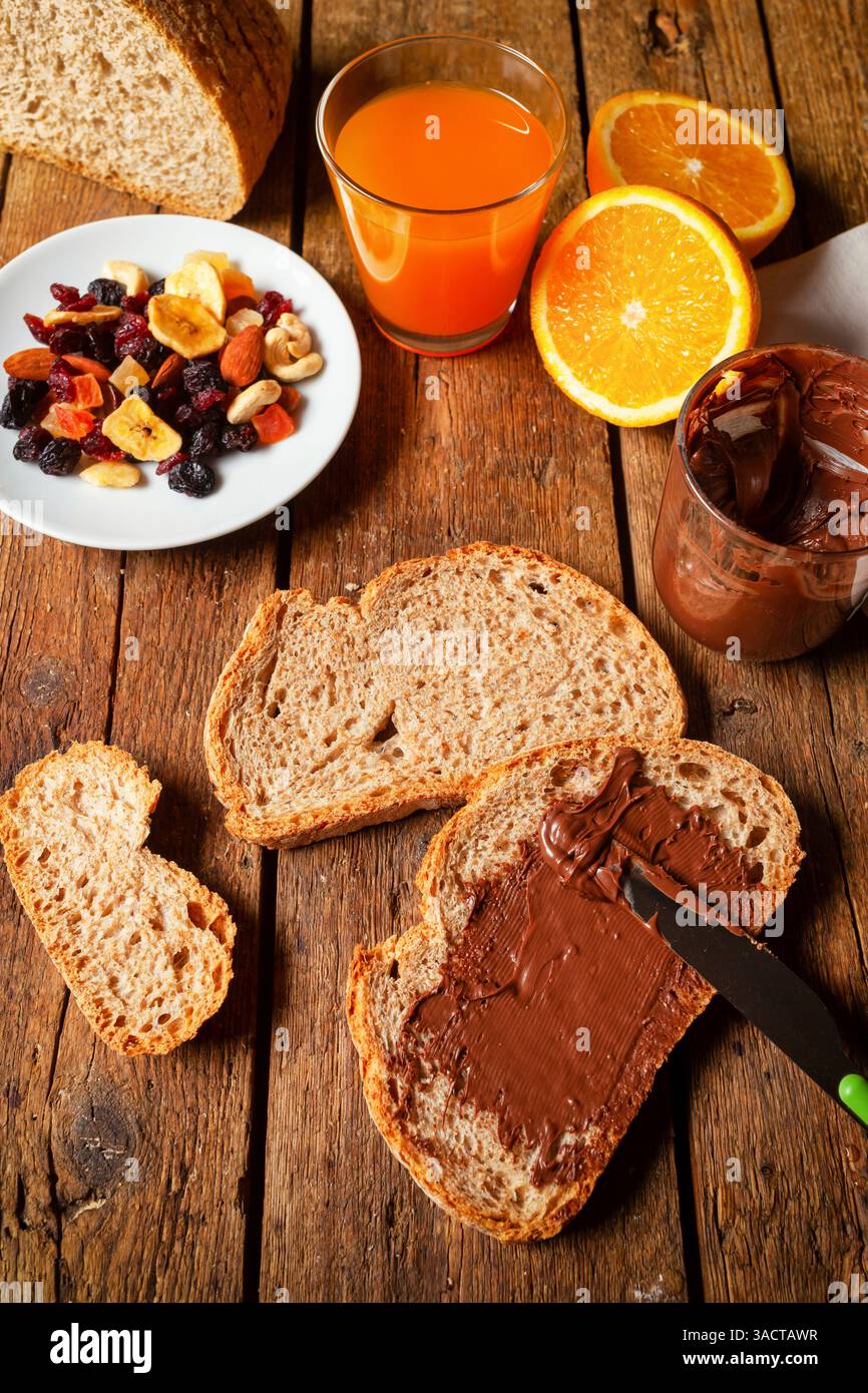 Fette di pane integrale con crema al cioccolato spalmata con coltello, bicchiere di succo d'arancia, frutta secca e arance su un tavolo rustico di legno per una colazione sana Foto Stock