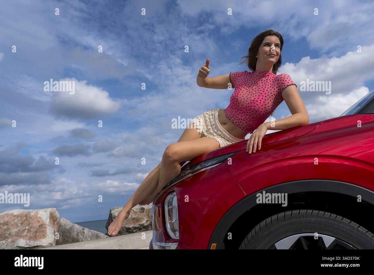 Una splendida modella rossa si siede con sicurezza sul cofano della sua auto, immergendosi nella brezza estiva mentre si imbarca in un'eccitante avventura di fondo, Foto Stock