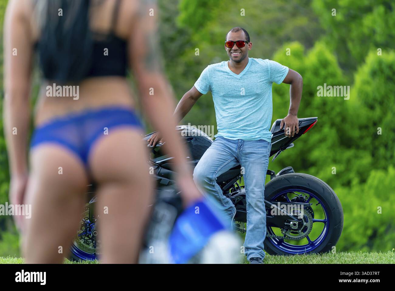 Una coppia multi-razziale posa amorevolmente accanto alla propria moto in un campo lussureggiante e verde. Abbracciati, sorridono con orgoglio, catturando entrambi i loro legami profondi Foto Stock